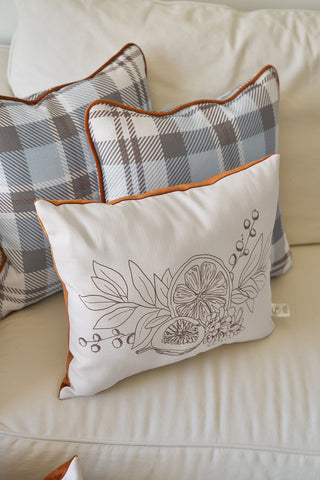 The Holiday Harvest Lumbar Pillow sits in front on a light sofa, featuring a botanical and citrus line drawing with orange piping, flanked by two blue and gray plaid pillows—an ideal autumnal accent.