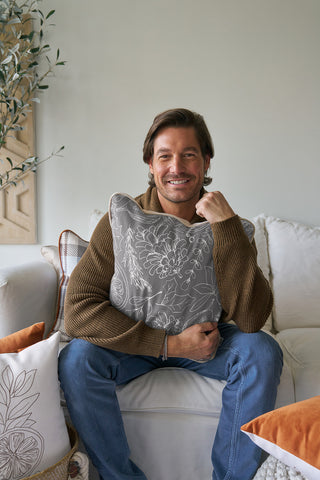A man in a brown sweater and jeans sits on a white sofa, smiling and hugging the Holiday Harvest Pillow. The Southern-style sofa also features decorative pillows, with a plant in the background.