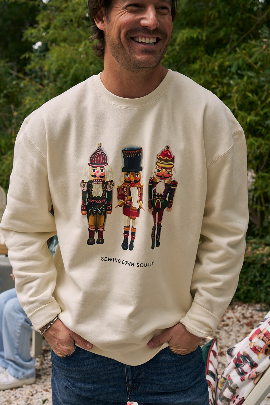 A smiling man wears the Nutcracker Crewneck, a cream festive sweatshirt featuring three colorful nutcrackers and SEWING DOWN SOUTH text. He stands outdoors with greenery in the background.