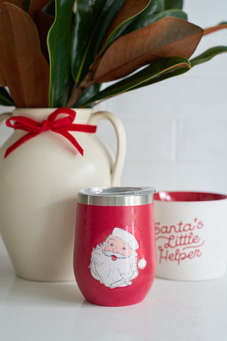 A Merry & Bright Tote + Santa Wine Tumbler Bundle features a red tumbler with a Santa Claus illustration, placed beside a white Santas Little Helper cup and a white vase adorned with a red ribbon and leafy branches.
