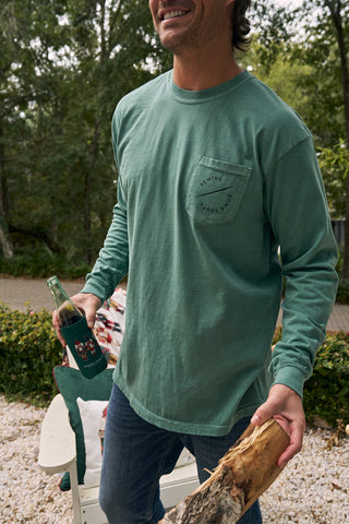 Wearing a Nutcracker Long Sleeve Tee and jeans, a person stands outdoors on a gravel path amid trees and greenery, holding a bottle in one hand and firewood in the other.