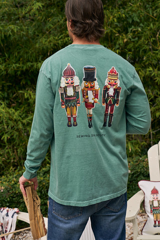 A person wearing a Nutcracker Long Sleeve Tee with three colorful nutcrackers on the back stands outdoors, holding firewood. Trees and white chairs in the background create a cozy, festive holiday atmosphere.