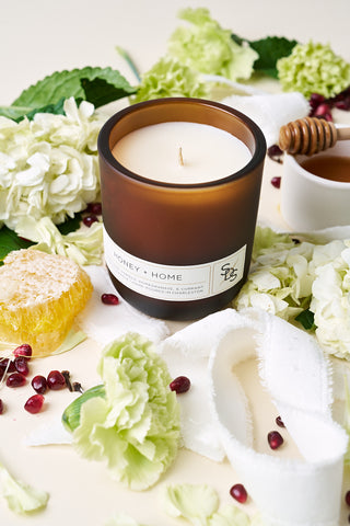 The HONEY + HOME 10 oz Candle in brown glass is set on a light surface, surrounded by green hydrangeas, pomegranate seeds, honeycomb, a honey dipper, and a white cloth.