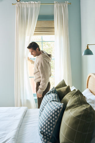 A man in a beige hoodie stands by a window with sheer curtains in a bedroom, looking outside. The bed in the foreground features green pillows and a Sage Stitch Grid Pillow; the walls are painted light blue.