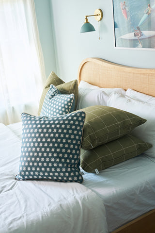 A neatly made bed features the Dimensional Dot in Sage Pillow stacked with green checkered pillows and a blue patterned pillow, beside a window with sheer curtains, wooden headboard, wall lamp, and surf-themed artwork above.