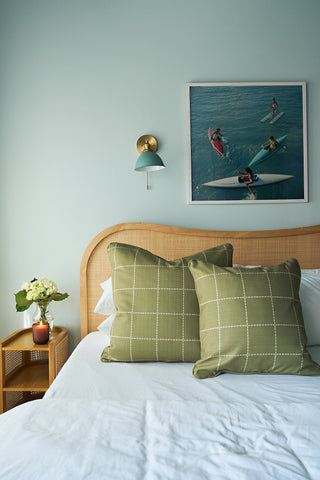 A neatly made bed features two Sage Stitch Grid Pillows in green plaid, a wooden headboard, a nightstand with a vase of flowers, and a framed photo of surfers above the bed. A wall sconce is mounted above the nightstand.