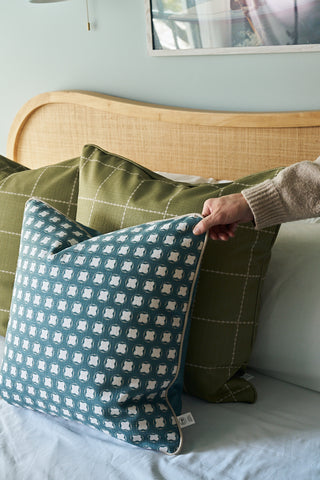 A person places the Dimensional Dot in Sage Pillow on a bed with two green grid-patterned pillows and a light wooden headboard.