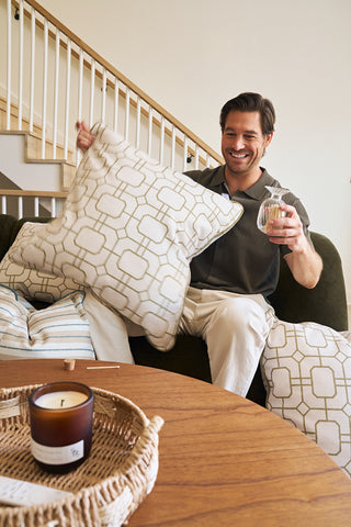 A smiling man sits on a couch holding a Sage Lattice Pillow in one hand and a drink in the other, surrounded by decorative pillows. A lit candle and a tray rest on the wooden table in front of him.