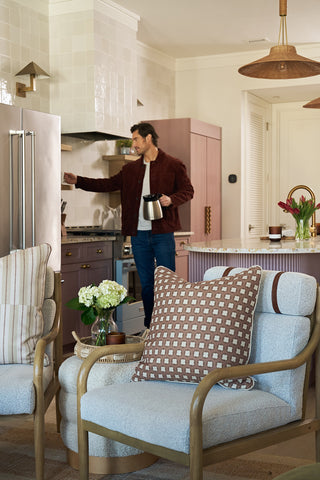 A man stands in a modern kitchen holding a coffee pot, reaching for the fridge. In front, two armchairs with Dimensional Dot in Rust Pillows and a small table with flowers create an inviting scene.