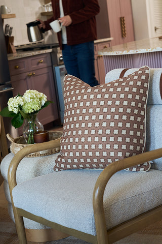 A cozy living space with a modern armchair featuring a Dimensional Dot in Rust Pillow, a side table holding a vase of white hydrangeas, and a person pouring a drink in the kitchen background.