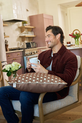 A smiling man sits on a chair in a cozy kitchen, holding a mug and phone. The Dimensional Dot in Rust Pillow rests on his legs, with flowers and kitchen items visible in the background.