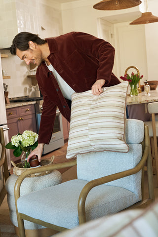 A man in a burgundy jacket smiles as he places the Rust King Street Stripes Pillow on a cushioned chair in a cozy, modern kitchen filled with flowers and natural light.