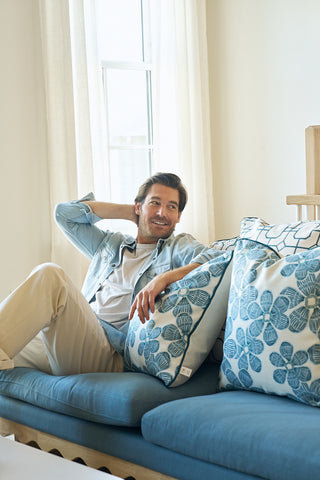 A man relaxes on a blue sofa with Broad Street Blooms in Navy pillows, smiling in a bright, sunlit room. The pillow covers are made in the USA from soft polyester.