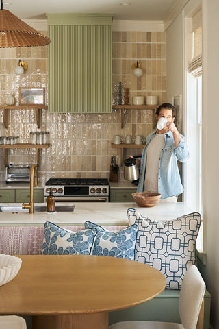 A person stands in a stylish kitchen holding a white mug and kettle. A Broad Street Blooms in Navy Pillow made in the USA accents the bench by a wooden table, with green cabinets, open shelves, and a tiled backsplash.