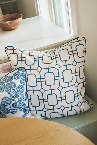 A large white pillow with a blue geometric square pattern sits next to the Navy Lattice Pillow, which features blue floral designs, on a light green bench by a window and a round wooden table.