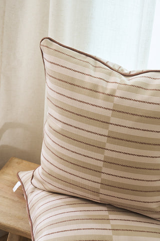 A close-up of two Rust King Street Stripes Pillows with beige and brown stripes, stacked on a light wooden stool in front of cream curtains, creating a cozy, minimal setting.