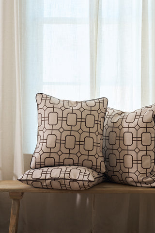A Rust Lattice Pillow adds a touch of coziness and modern style as it sits on a wooden bench in front of sheer white curtains and a window, its geometric pattern enhancing the inviting atmosphere.