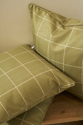 Two Sage Stitch Grid Pillows in green with a white grid pattern rest on a wooden surface, leaning against a light-colored wall.