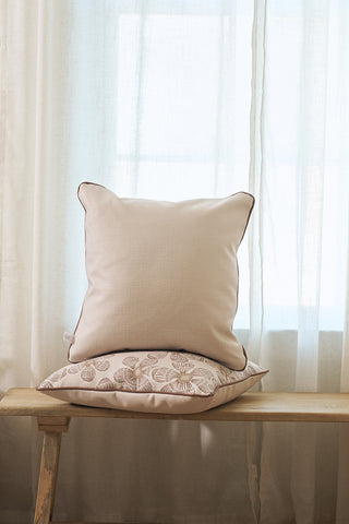 The Broad Street Blooms in Rust Pillow, featuring a light hue with dark piping and a discreet zipper, rests upright atop another patterned pillow on a wooden bench before sheer curtains and a sunlit window.