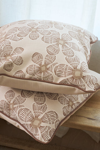 Two Broad Street Blooms in Rust Pillows, featuring beige covers with brown abstract floral patterns and zipper closures, are stacked on a wooden bench before a white curtain, creating a cozy and inviting atmosphere.