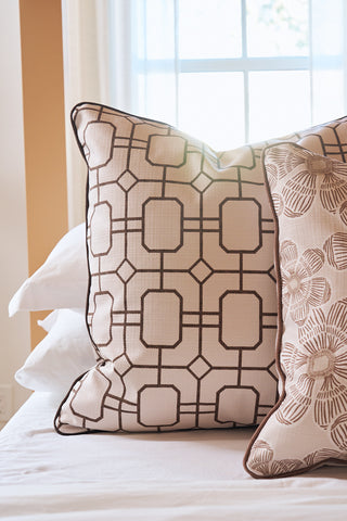 Two decorative pillows rest on a bed in front of a bright window. One is the Rust Lattice Pillow, featuring a geometric square and diamond pattern; the other shows a floral design. White bedsheets are visible underneath.