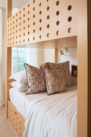 A modern wooden bunk bed with circular cutouts features a neatly made lower bed with white linens and a Rust Lattice Pillow, positioned by a window draped with sheer curtains.