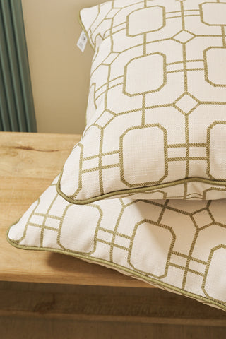 Two Sage Lattice Pillows, featuring a green geometric pattern on white, are stacked on a light wood surface in front of a neutral wall and vertical green radiator.