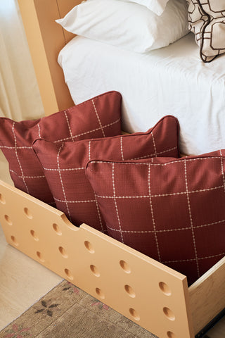 A beige under-bed storage drawer holds three Rust Stitch Grid Pillows. The bed, dressed in white linens, is beside a patterned cushion, with part of a rug visible on the floor.