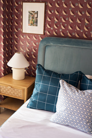 A cozy bedroom corner with a blue upholstered bed, the Navy Stitch Grid Pillow, a wooden nightstand topped with a white lamp, and red bird-patterned wallpaper. A framed picture hangs above the nightstand.