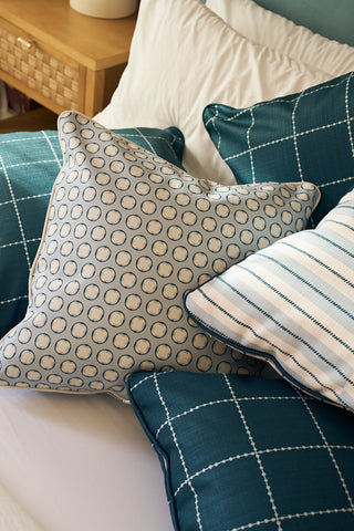 A close-up of a bed with the Dimensional Dot in Navy Pillow, featuring geometric patterns in teal and blue tones, arranged against crisp white bedding.