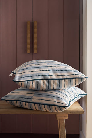Two Navy King Street Stripes euro pillow covers, made in the USA, are stacked on a wooden bench in front of a mauve cabinet with vertical panels and wooden handles. Light shines from the right side.