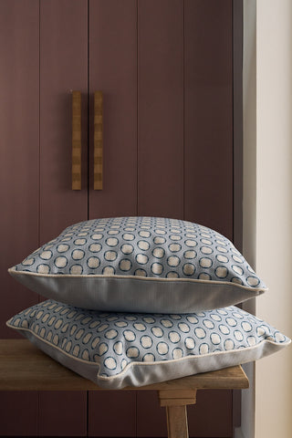 Two Dimensional Dot in Navy Pillows, featuring a light gray base with blue and white circle patterns, are stacked on a wooden bench. Behind them stands a mauve cabinet with vertical wooden handles.