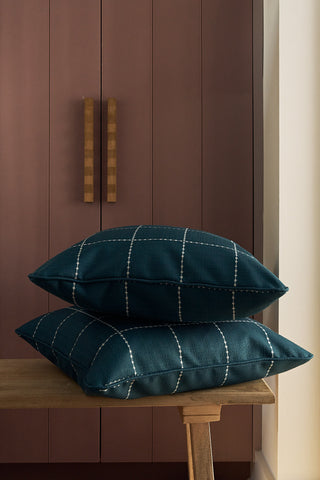 Two Navy Stitch Grid Pillows are stacked on a wooden bench in front of a mauve-colored cabinet with wooden handles.