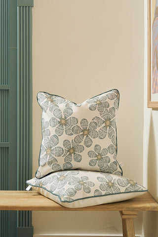 Two decorative pillows with a gray and yellow floral pattern, including a 19x19 cover, are stacked on a wooden bench against a light wall beside the Broad Street Blooms in Sage Pillow cover and framed artwork.