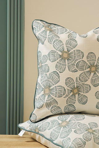 A close-up of two stacked cushions, including the Broad Street Blooms in Sage Pillow with a cream background and teal floral pattern, placed near a teal vertical-groove wall on a light wooden surface.