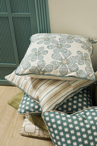 The Broad Street Blooms in Sage Pillow, featuring floral, plaid, and geometric patterns in teal, green, and cream, is stacked with other decorative covers on a light wooden floor beside a teal panel wall.