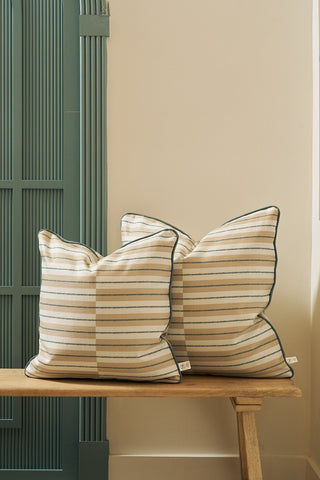 Two Sage King Street Stripes Pillow covers in beige with dark green stripes and trim are stacked on a light wooden bench, positioned in front of a cream wall and teal vertically paneled room divider.