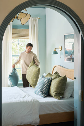 A man arranges decorative Sage Stitch Grid Pillows on a neatly made bed with a wooden frame in a room featuring soft blue walls, white-curtained large window, modern wall sconce, and an arched doorway framing the scene.
