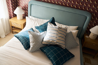 A neatly made bed with a light blue upholstered headboard is piled with patterned pillows, including the Navy King Street Stripes Pillow. The room has red wallpaper with white birds, a wooden nightstand, lamp, framed picture, and sheer curtains.