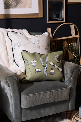 A cozy armchair with white fabric is topped with a modern accent pillow and the Winter Sports Gear Lumbar Pillow, featuring ski equipment illustrations, adding warmth and style to the room.