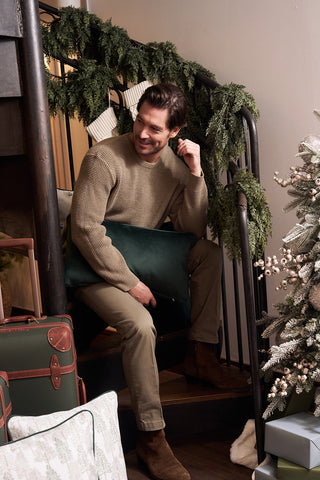 A man in a beige sweater sits on garland-decorated stairs, smiling and holding a green holiday pillow. Next to him are luggage and festive pillows, including the Row of Trees Extended Lumbar Pillow, by a decorated Christmas tree.