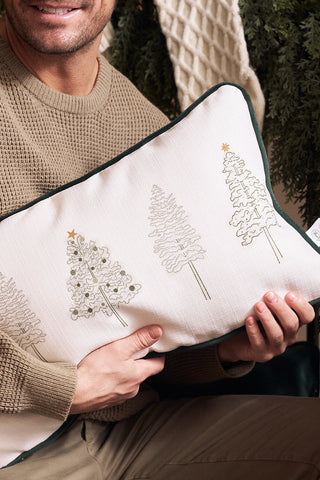 A person in a tan sweater holds the Row of Trees Extended Lumbar Pillow, featuring embroidered Christmas trees and gold stars. Greenery and a knit blanket are visible in the background.