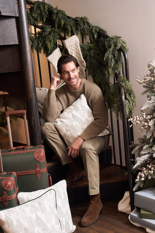 A man sits on garland-decorated stairs, smiling and holding a Christmas Tree Farm Pillow. Suitcases and more Christmas Tree Farm Pillows are beside him, with a decorated Christmas tree partially visible on the right.