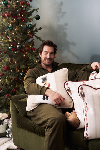 A man in green pajamas sits on a green sofa, smiling and holding a Nutcracker Stripe Pillow in Angora. Beside him, a decorated Christmas tree with red and green ornaments and collectible Nutcrackers creates festive charm.