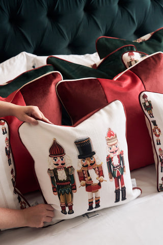 A person arranges festive pillows on a bed, including a holiday decorative pillow, a wine velvet pillow, and the Nutcracker Holiday Pillow adorned with nutcracker illustrations.