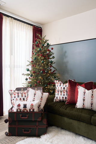 A decorated Christmas tree stands by a window with sheer curtains. A green couch with Sewing Down South holiday velvet pillows and Nutcracker Line Up Pillows sits beside vintage suitcases stacked with more Nutcracker Line Up Pillows. A fluffy white rug covers the floor.