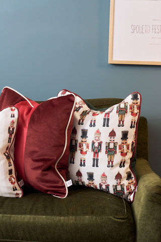 Two decorative pillows rest on a green sofa: a Red Blocking Velvet Pillow and a festive holiday pillow with colorful nutcracker figures. The blue wall and framed artwork in the background add extra charm to the scene.