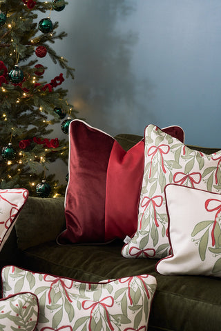 A green velvet sofa is decorated with the Red Blocking Velvet Pillow and festive holiday pillows, such as white ones featuring green and red bows. In the background, a lit Christmas tree sparkles with ornaments.
