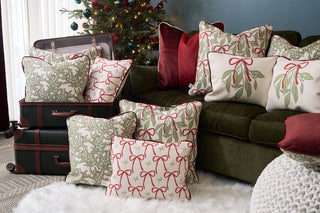 The Jolly Holly Pillow from Sewing Down South is arranged on a green sofa and stacked suitcases near a Christmas tree, creating a cozy, festive holiday atmosphere.