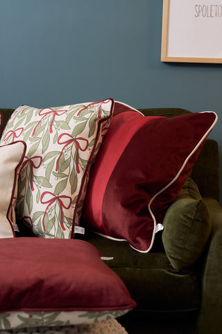 A dark green sofa with decorative cushions, including a festive holiday pillow with a green and red ribbon pattern and the Red Blocking Velvet Pillow, set against a blue wall with partially visible framed artwork.